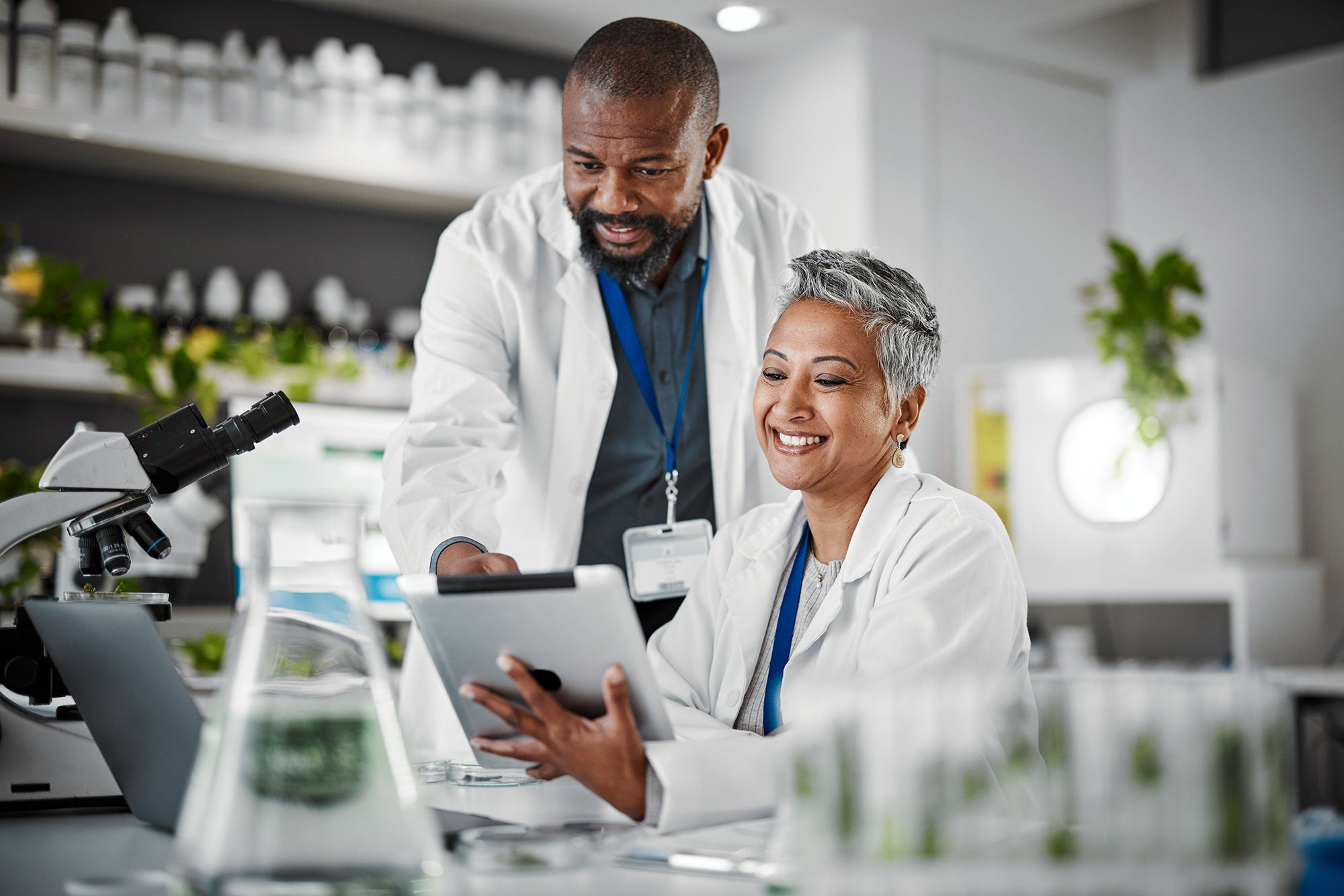Two scientists in a lab setting, one using a tablet and the other observing, with laboratory equipment around.