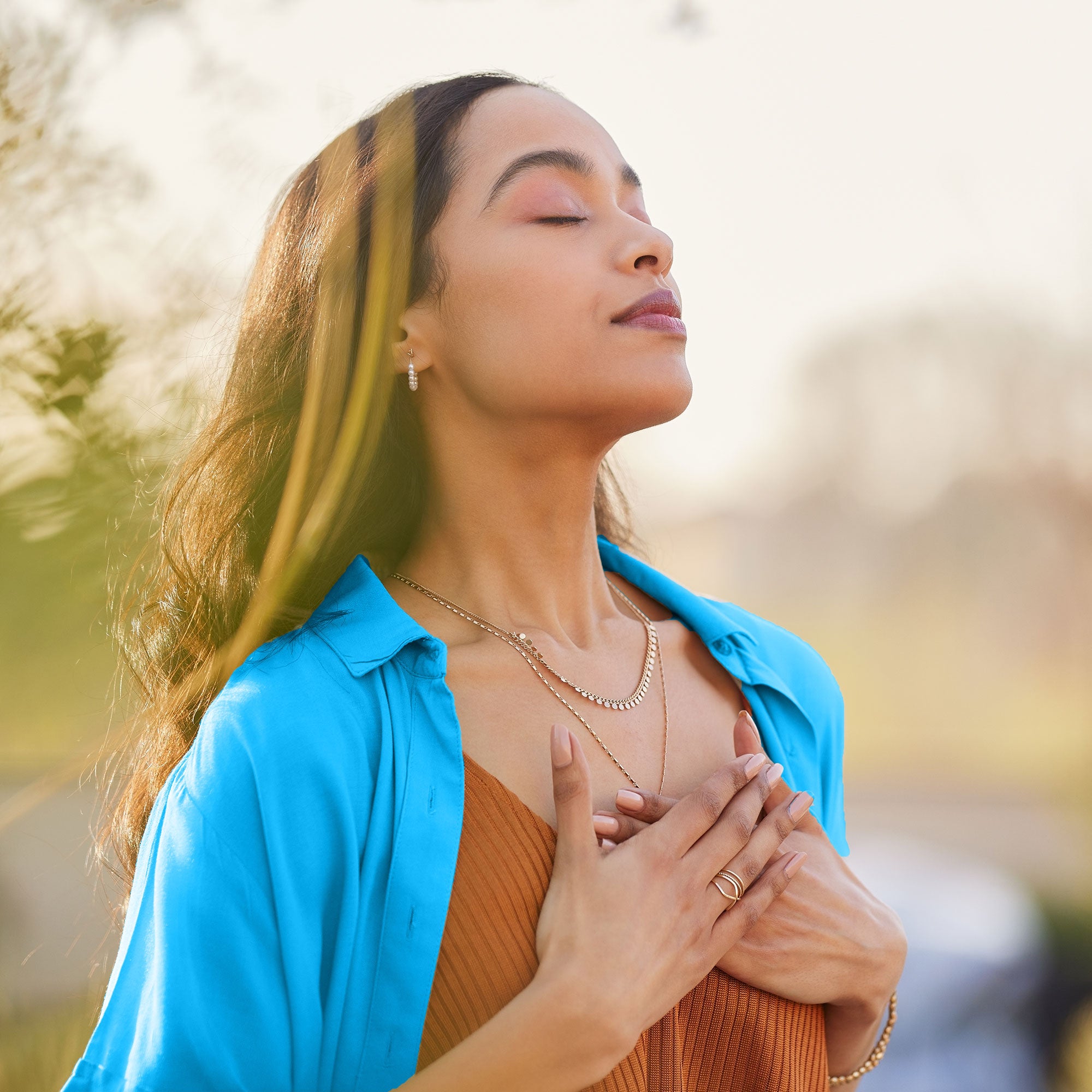 Woman in a blue shirt and brown top with hands over her heart, outdoors.
