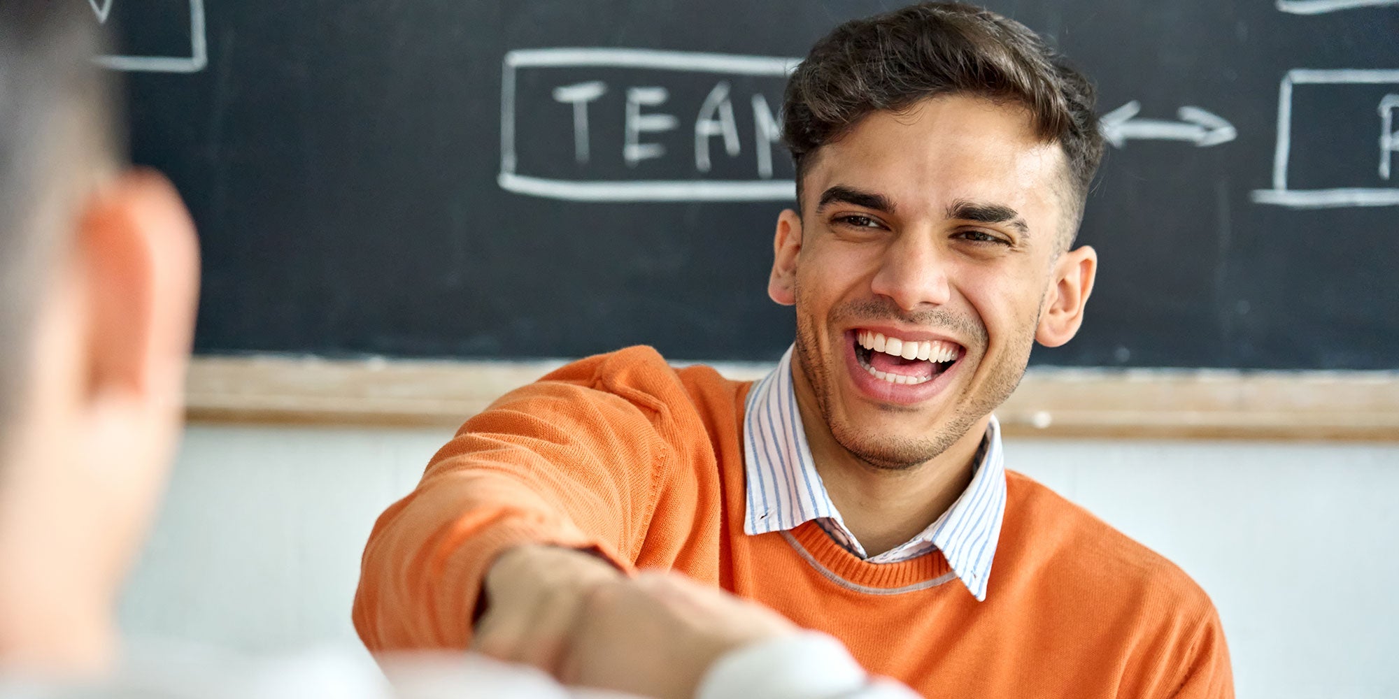 Man in an orange sweater shaking hands with another person in a classroom setting.