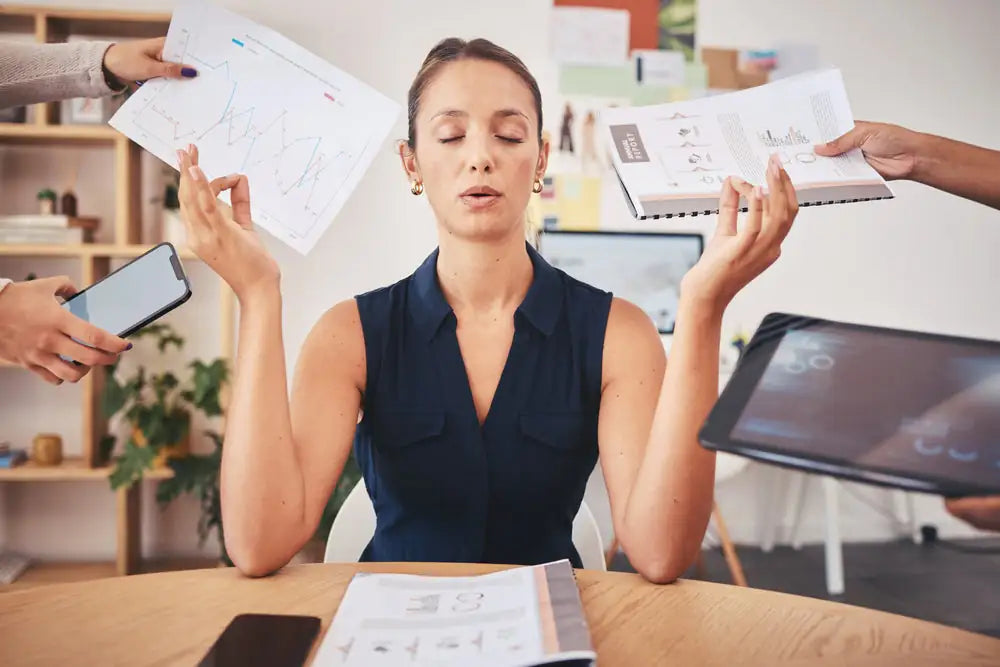 A woman sitting at her desk with her eyes closed staying calm surrounded by people's hands holding lots of work elements such as a report, a phone, a laptop to show stress at work.