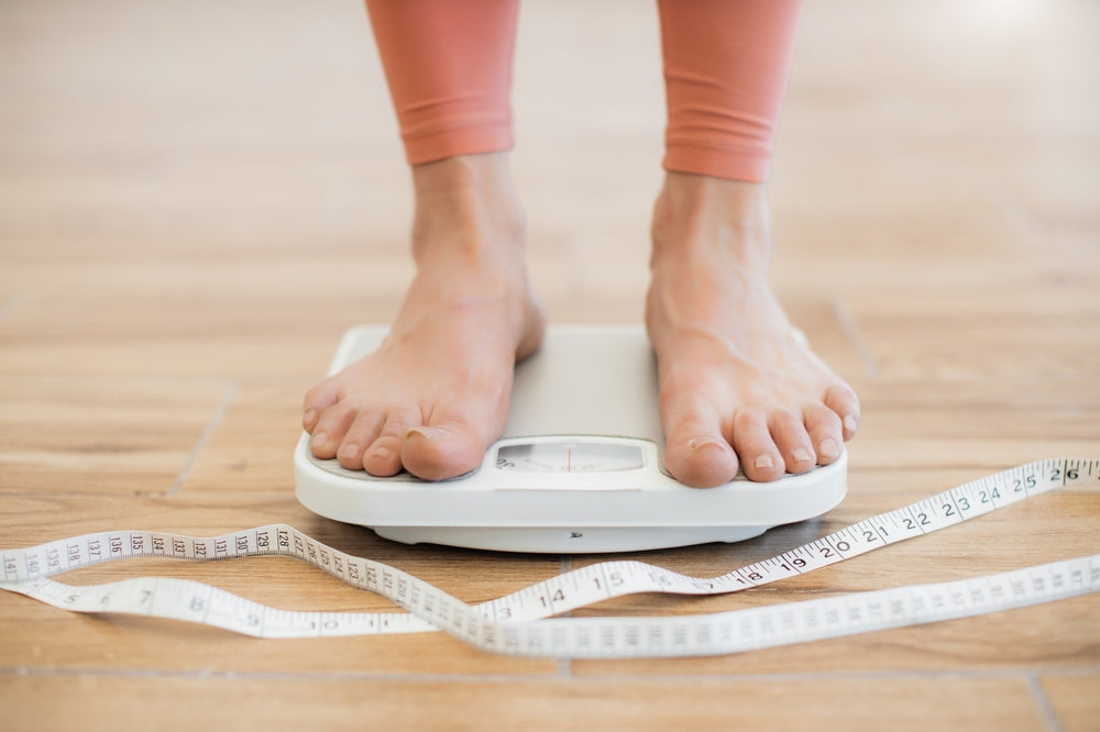 CLose up of a woman's feet standing on a set of scales with a measuring tape on the ground to represent weight management