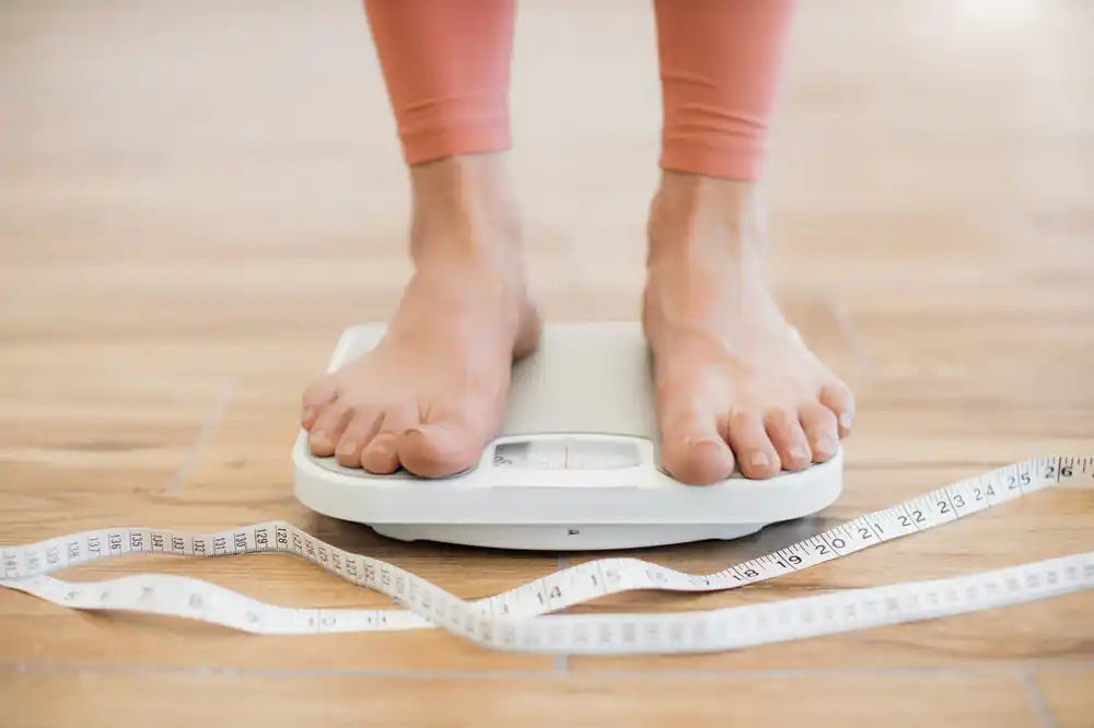 CLose up of a woman's feet standing on a set of scales with a measuring tape on the ground to represent weight management