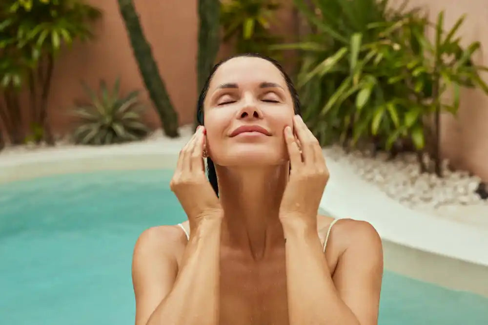 A woman in an outdoor pool, with her finger tips on her face, smiling with her eyes closed enjoying her holiday.