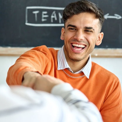 Man in orange sweater smiling and shaking hands with another person in front of a blackboard with 'TEAM' written on it.