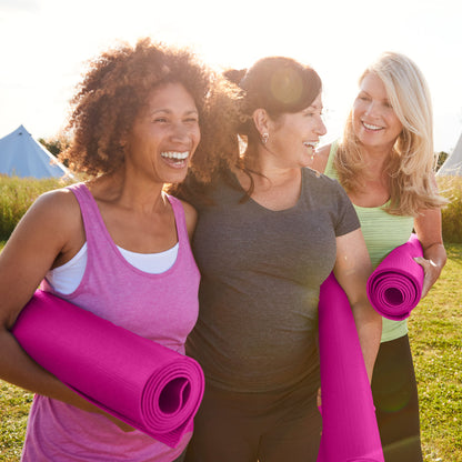 Three women holding pink yoga mats outdoors on a sunny day.